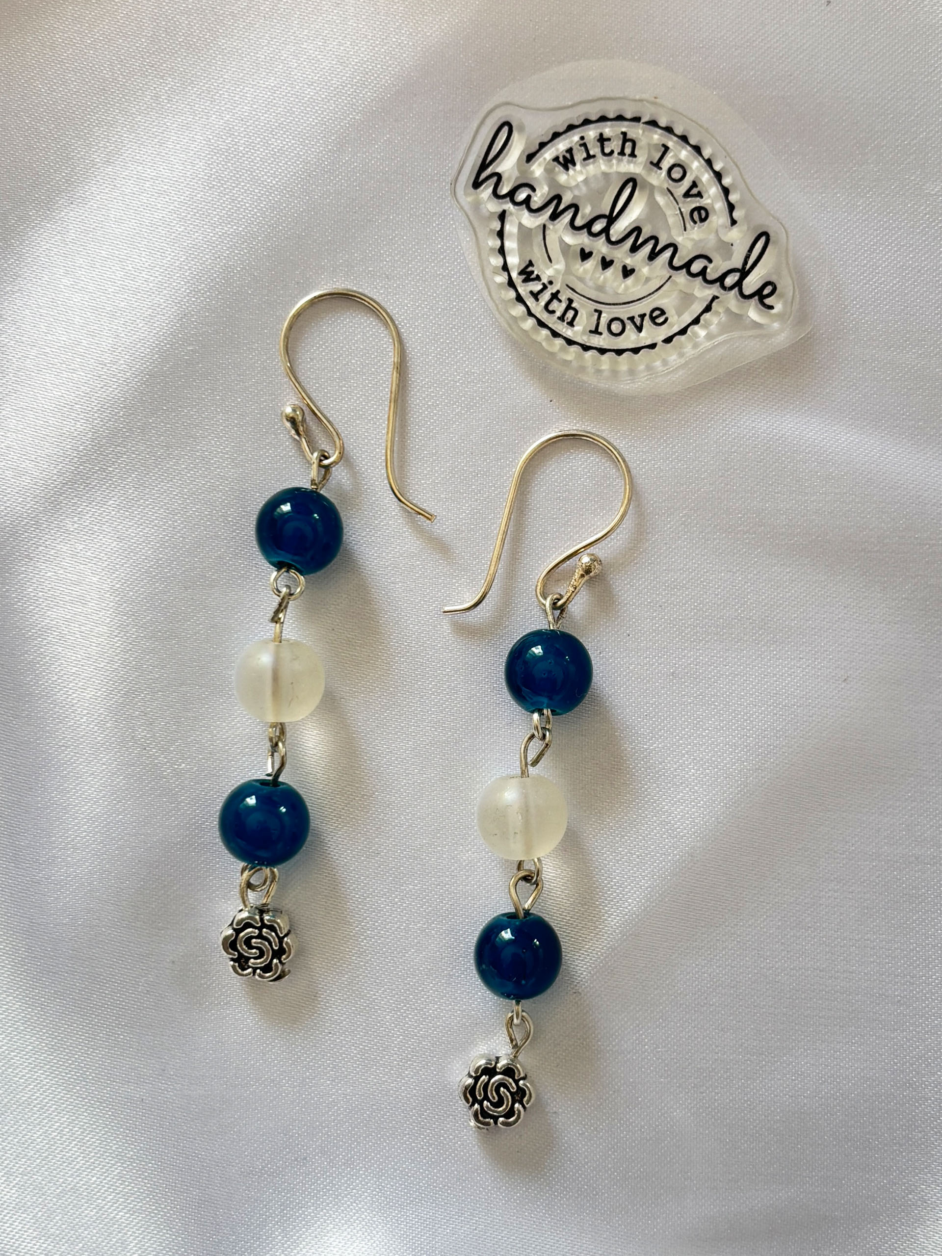 Ocean Bloom Drop Earrings - These graceful drop earrings feature deep teal blue beads paired with soft translucent accents, finished with a delicate floral charm at the base. The cascading design adds movement and charm, making them perfect for both everyday wear and special occasions.